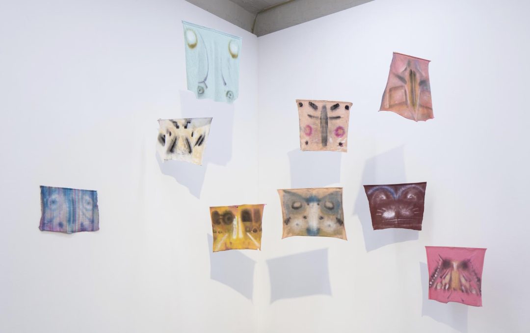 A series of nine mono prints on t-shirt fabric hang mid air in a mobile formation. Like mythical death masks, the monotypes reflect this process of selection and appropriation. They appear as Rorschach or butterfly prints.